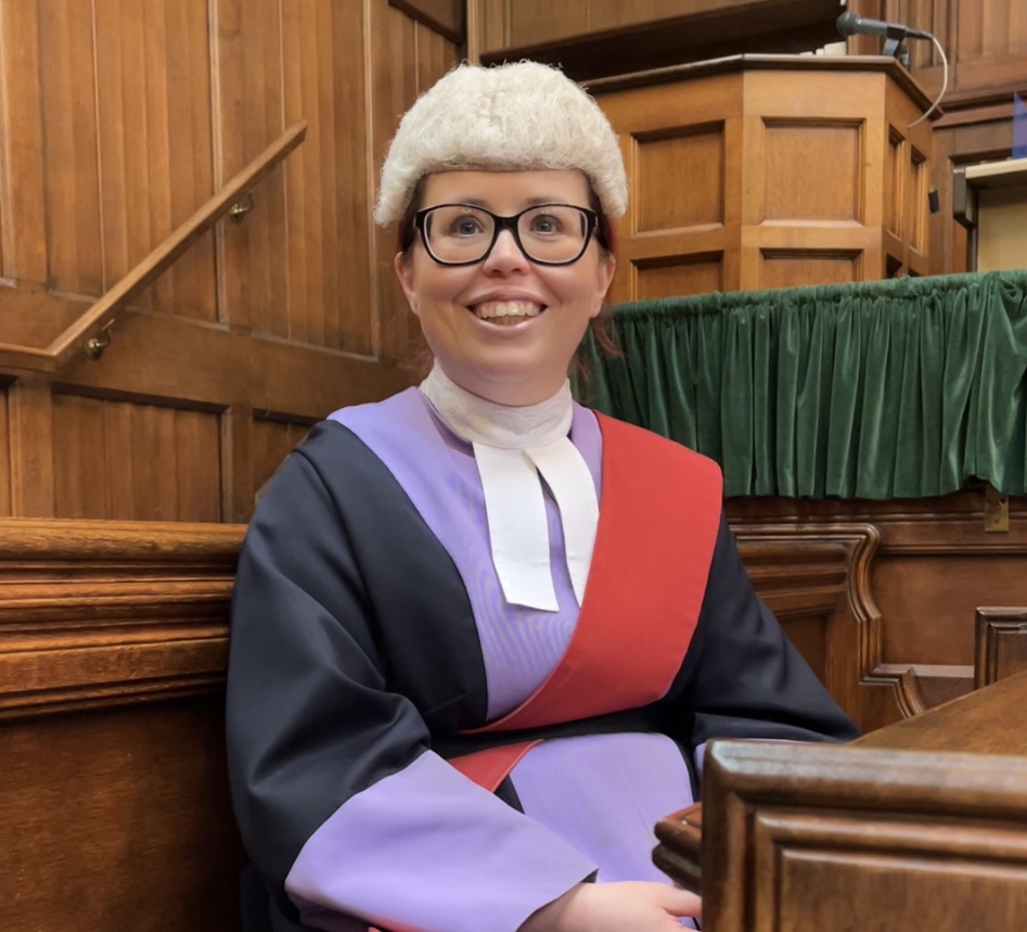 Louise smiling wearing a wig and a black gown with purple sleeves and a purple and red collar. 